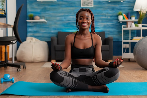 Colocar mujer negra practicando yoga sentada con las piernas cruzadas en top deportivo y leggins en posición de loto. Practicando la armonía de la mente tranquila para una vida sin estrés en la sala de estar del hogar.