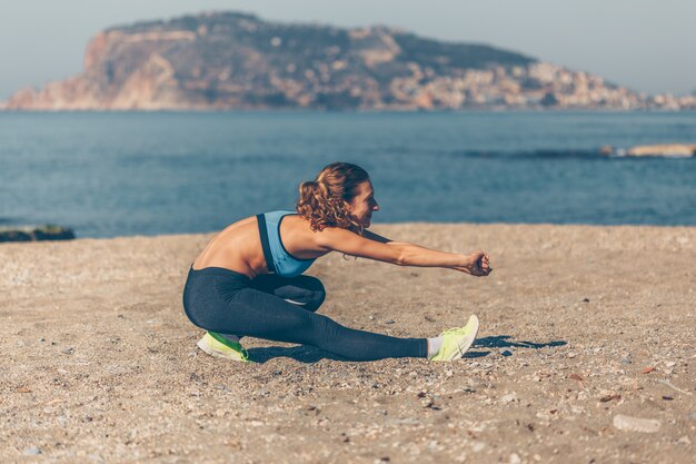 Colocar mujer en ropa deportiva haciendo ejercicio de calentamiento en la playa durante el día con mar
