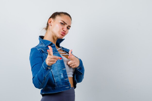 Colocar mujer en top corto, chaqueta de jean, leggings apuntando a la cámara con los dedos índices y mirando confiado, vista frontal.