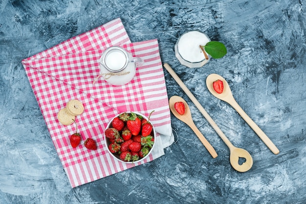 Coloque un tazón de fresas y una jarra de leche sobre una toalla de cuadros rojos con cucharas de madera y un tazón de vidrio con yogur sobre una superficie de mármol azul oscuro. horizontal