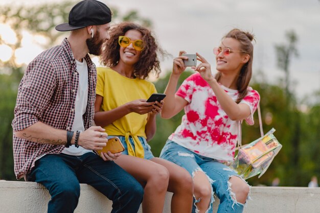 Colorida y elegante compañía joven feliz de amigos sentados en el parque, hombres y mujeres que se divierten juntos