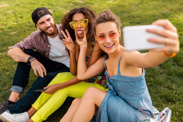 Colorida y elegante compañía joven feliz de amigos sentados en el parque, hombres y mujeres que se divierten juntos