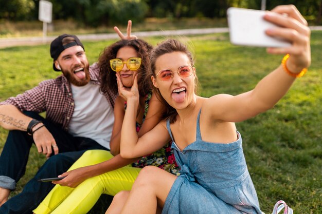 Colorida y elegante compañía joven feliz de amigos sentados en el parque, hombres y mujeres que se divierten juntos