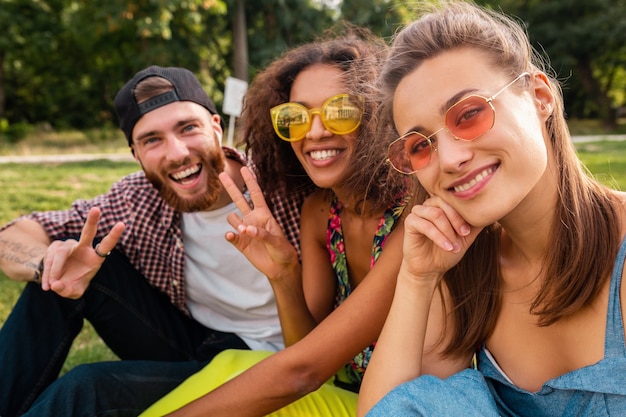 Colorida y elegante compañía joven feliz de amigos sentados en el parque, hombres y mujeres que se divierten juntos