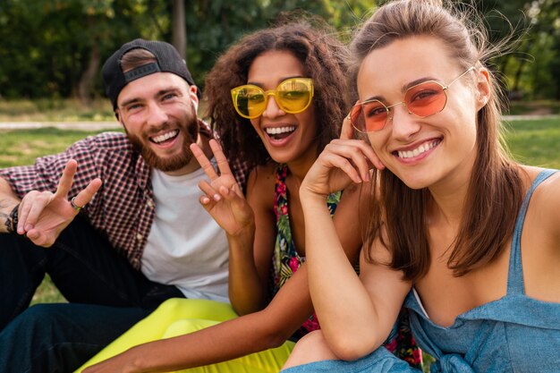 Colorida y elegante compañía joven feliz de amigos sentados en el parque, hombres y mujeres que se divierten juntos