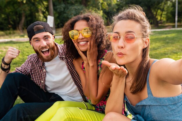 Colorida y elegante compañía joven feliz de amigos sentados en el parque, hombres y mujeres que se divierten juntos