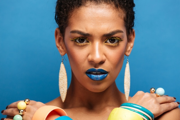 Colorida y estricta mujer seria de raza mixta con maquillaje de moda y accesorios posando con las manos cruzadas sobre los hombros, sobre la pared azul