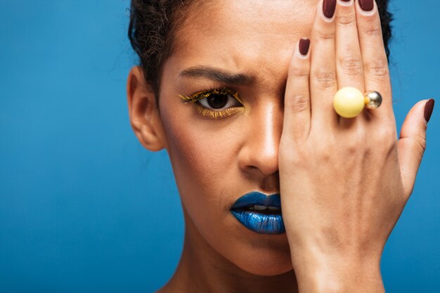 Colorida mujer asustada u ofendida de raza mixta con maquillaje moderno y accesorios que cubren los ojos con la mano, sobre la pared azul