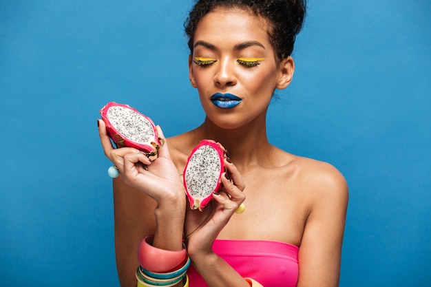 Foto gratuita colorida mujer de raza mixta bonita con cosméticos brillantes en la cara con pitahaya madura cortada por la mitad en ambas manos aisladas, sobre pared azul