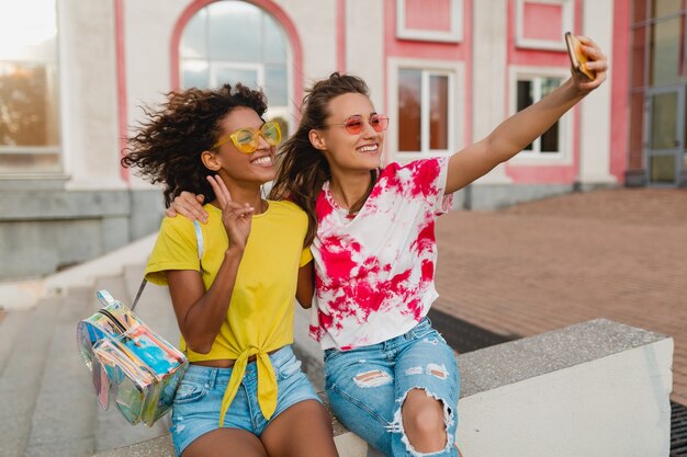 Colorido retrato de amigos de niñas jóvenes felices sonriendo sentado en la calle tomando fotos selfie en teléfono móvil, mujeres divirtiéndose juntos