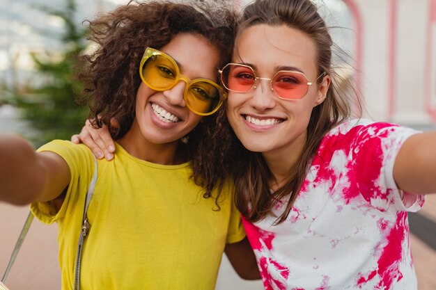 Colorido retrato de amigos de niñas jóvenes felices sonriendo sentado en la calle tomando fotos selfie en teléfono móvil, mujeres divirtiéndose juntos