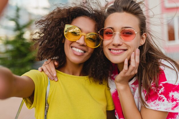 Colorido retrato de amigos de niñas jóvenes felices sonriendo sentado en la calle tomando fotos selfie en teléfono móvil, mujeres divirtiéndose juntos