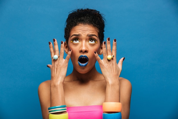 Colorido retrato de mujer mulata con maquillaje elegante emocionalmente demostrando joyas en las manos mirando hacia arriba, sobre la pared azul