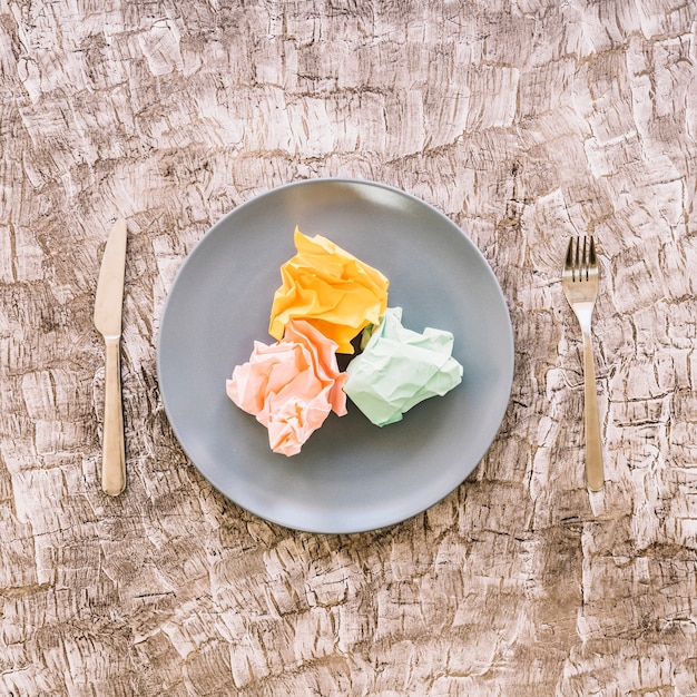 Foto gratuita coloridos papeles arrugados en placa entre tenedor y cuchillo de cocina sobre superficie de madera