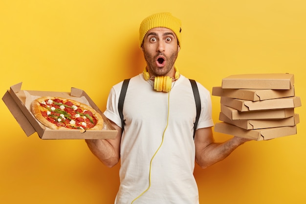 El comerciante de pizzas tiene cajas de cartón con bocadillos, se ve con expresión omg, usa un sombrero amarillo y una camiseta blanca, impresionado por algo, tiene mucho trabajo