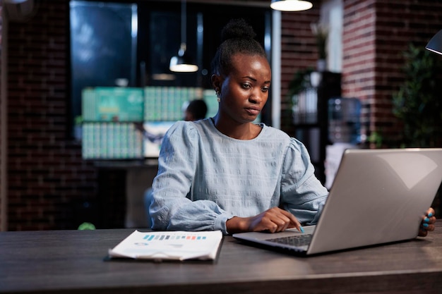 Comerciante profesional del mercado de valores de Forex sentado en el escritorio con una computadora portátil mientras compra y vende opciones comerciales. Mujer afroamericana sentada en el espacio de trabajo de la oficina mientras analiza datos financieros en tiempo real