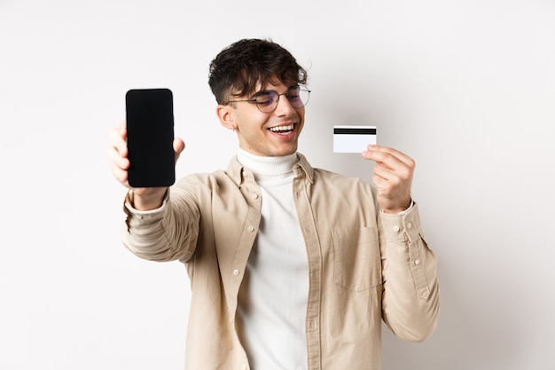 Comercio electrónico feliz joven mirando satisfecho con tarjeta de crédito de plástico que muestra la pantalla del teléfono inteligente a boas ...