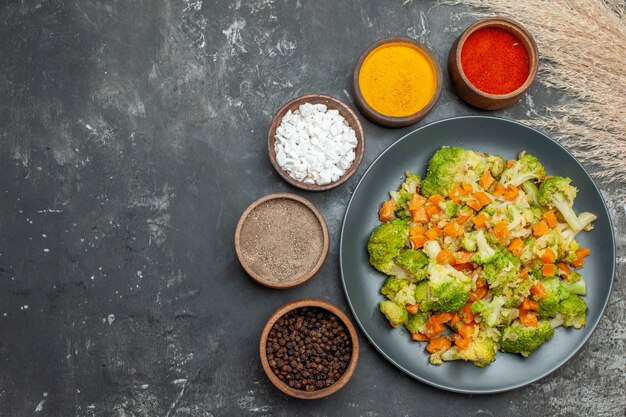 Comida saludable con brocoli y zanahorias en un plato negro y especias en mesa gris