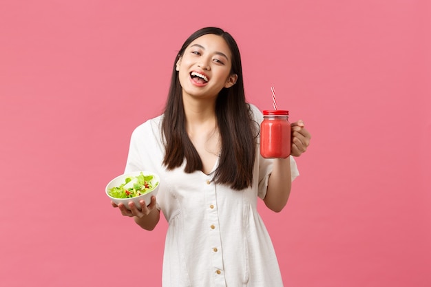 Comida sana, emociones y concepto de estilo de vida de verano. Entusiasta y optimista linda chica asiática llena de energía, comiendo sabrosa ensalada fresca y bebiendo batidos, sonriendo a la cámara con fondo rosa feliz.