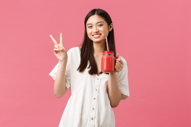 Comida sana, emociones y concepto de estilo de vida de verano. Feliz chica asiática guapa con vestido blanco, mostrando el signo de la paz kawaii y bebiendo batido de vidrio y paja.