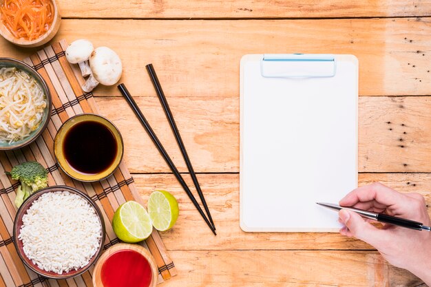 Comida tailandesa con la mano de una persona escribiendo en el portapapeles con lápiz en el escritorio de madera