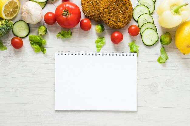 Comida vegana junto al cuaderno vacío en la mesa de madera blanca