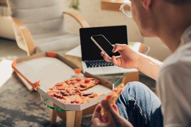 Comiendo pizza. Hombre estudiando en casa durante cursos en línea, escuela inteligente. Obtener clases o profesión mientras está aislado, cuarentena contra la propagación del coronavirus. Usando computadora portátil, teléfono inteligente, auriculares.