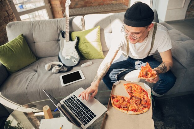 Comiendo pizza. Hombre estudiando en casa durante cursos en línea, escuela inteligente.