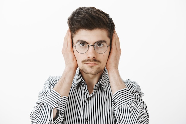 Compañero de cuarto ruidoso distrae al chico del trabajo independiente. Retrato de un compañero de trabajo masculino europeo común molesto molesto en gafas de moda y camisa a rayas, cubriendo las orejas con las palmas, mirando seriamente