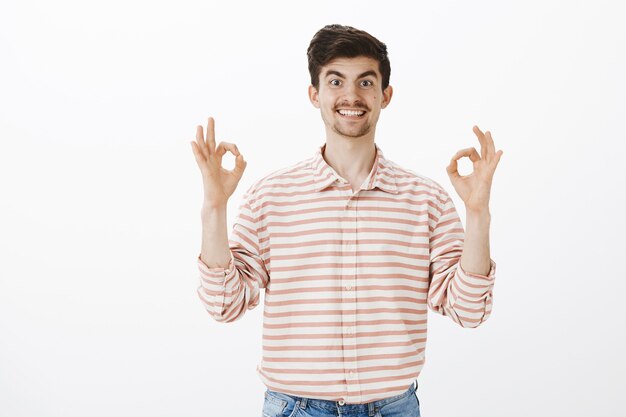 Compañero de trabajo útil dispuesto a ayudar. Retrato de un hombre europeo amistoso enérgico con bigote, levantando las manos y mostrando una buena o buena señal, aprobando la idea y siendo feliz de asegurar un amigo sobre la pared gris