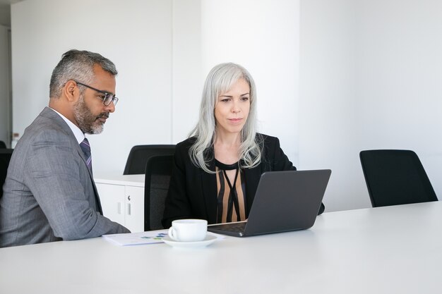 Compañeros amigables que usan la computadora portátil para videollamadas, sentados a la mesa con una taza de café, mirando la pantalla y hablando. Concepto de comunicación online