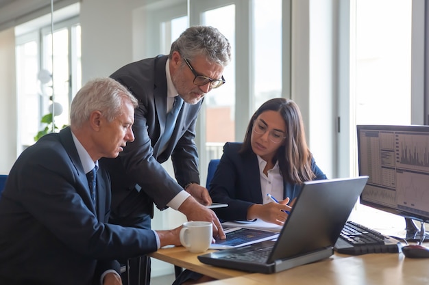 Foto gratuita compañeros concentrados mirando gráficos estadísticos y hablando sobre el trabajo. altos directivos profesionales y joven asistente preparando plan de negocios. concepto de trabajo en equipo, gestión y asociación