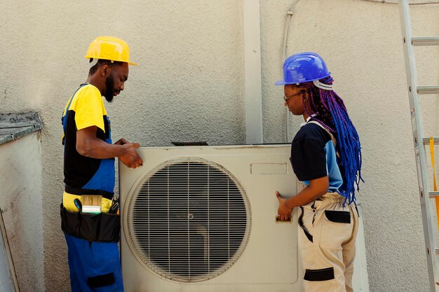 Los compañeros de trabajo afroamericanos de ingeniería abren un acondicionador de aire fuera de servicio oxidado para reemplazarlo con un nuevo condensador exterior de alto rendimiento después de drenar el refrigerante y reemplazar los conductos
