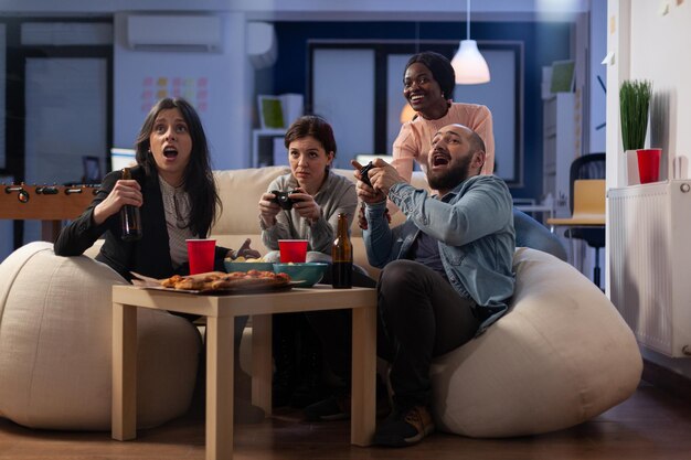 Compañeros de trabajo alegres celebrando la victoria de los videojuegos en la fiesta de la oficina, divirtiéndose con bebidas y bocadillos después de las horas de trabajo. Compañeros de trabajo jugando con joystick, reuniéndose para beber cerveza y disfrutar de la celebración.