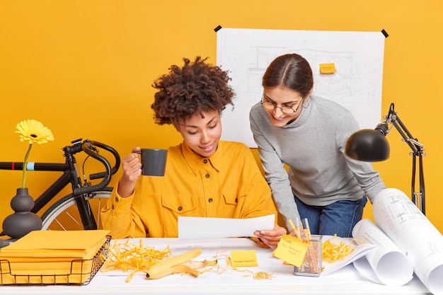 Compañeros de trabajo alegres de raza mixta discuten ideas para proyectos, comparten opiniones entre sí, se preparan para la presentación de bocetos arquitectónicos para la nueva pose de construcción en el escritorio con papeles alrededor