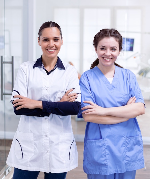 Compañeros de trabajo dentistiry felices sonriendo de pie en línea Retrato del equipo de estomatología en la recepción dental con los brazos cruzados mirando a la cámara con uniforme.