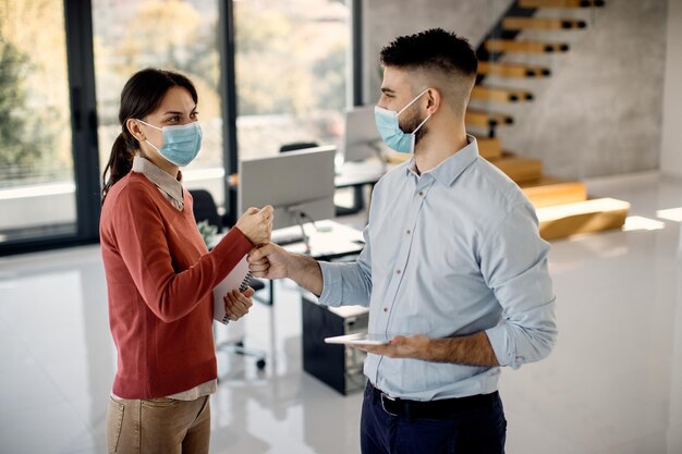 Compañeros de trabajo felices con máscaras faciales mientras saludan con los puños en la oficina