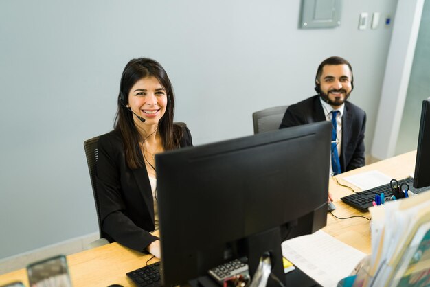 Compañeros de trabajo felices sonriendo y haciendo contacto visual mientras están sentados en su escritorio en un centro de llamadas. Colegas ejecutivos esperando recibir llamadas de clientes para soporte técnico
