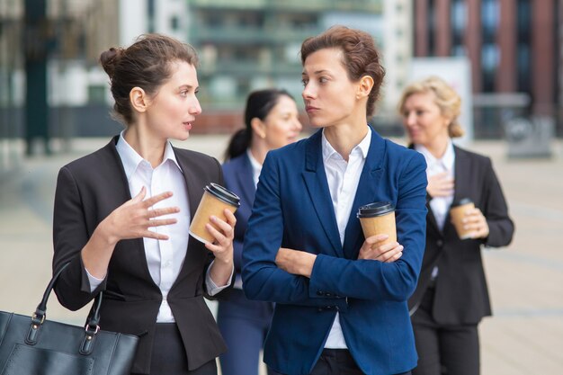 Compañeros de trabajo femeninos con tazas de café para llevar caminando juntos en la ciudad, hablando, discutiendo proyectos o charlando. Tiro medio. Concepto de descanso laboral