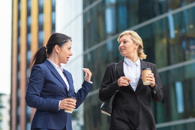 Compañeros de trabajo jóvenes y de mediana edad con tazas de café para llevar caminando juntos al aire libre, hablando, discutiendo proyectos o charlando. Tiro medio. Concepto de descanso laboral