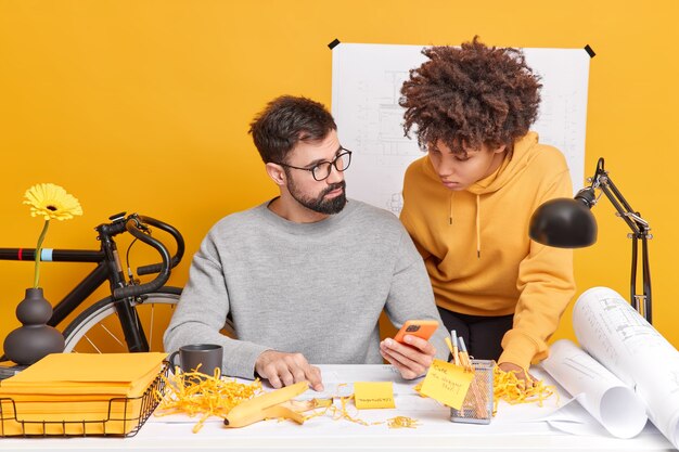 Compañeros de trabajo de mujer y hombre de raza mixta intentan encontrar una solución de información de verificación de trabajo en Internet a través de los documentos de estudio de teléfonos móviles que se plantean en el espacio de coworking disfrutan de la cooperación