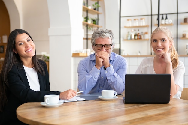 Compañeros de trabajo o socios de diferentes edades reunidos con una taza de café en el coworking, sentados a la mesa con un portátil y documentos,