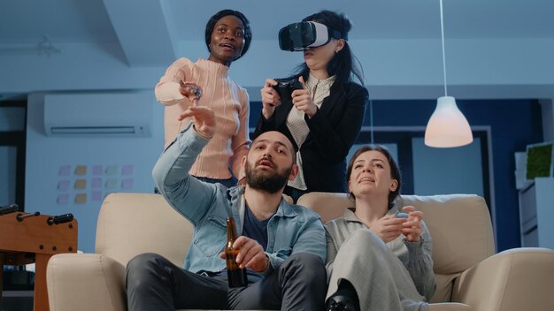 Compañeros de trabajo que usan tecnología para jugar videojuegos después del trabajo. Colegas jugando con gafas vr y controlador en televisión, disfrutando de bebidas y bocadillos después de horas de diversión.