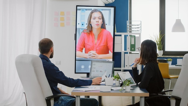 Compañeros de trabajo que utilizan videollamadas en la televisión para conversar con una mujer sobre un proyecto empresarial. Compañeros hablando con el gerente en una conferencia en línea para informar la estrategia sobre la presentación