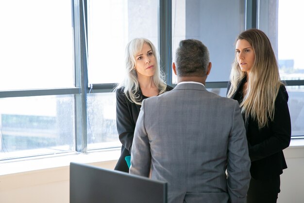 Compañeros de trabajo serios hablando con el jefe masculino juntos, de pie en la oficina, discutiendo el proyecto. Plano medio, vista trasera. Concepto de reunión de grupo o comunicación empresarial