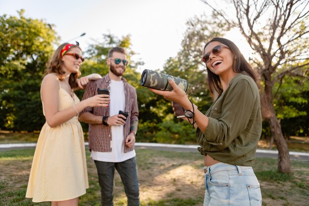 Compañía joven inconformista de amigos que se divierten juntos en el parque sonriendo escuchando música en el altavoz inalámbrico, temporada de verano