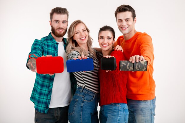 Compañía joven inconformista de amigos que se divierten juntos sonriendo escuchando música en altavoces inalámbricos, fondo blanco de estudio aislado en traje elegante colorido, mostrando dispositivos en la cámara