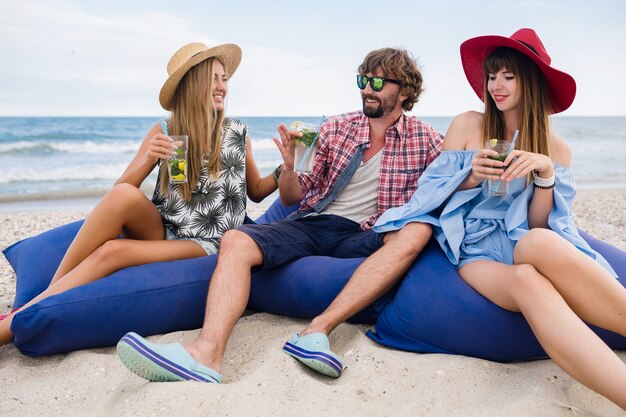 Compañía joven inconformista de amigos de vacaciones en el café de la playa, bebiendo un cóctel mojito, feliz positivo, estilo veraniego, sonriendo feliz, dos mujeres y un hombre divirtiéndose juntos, hablando, coqueteando, romance, tres