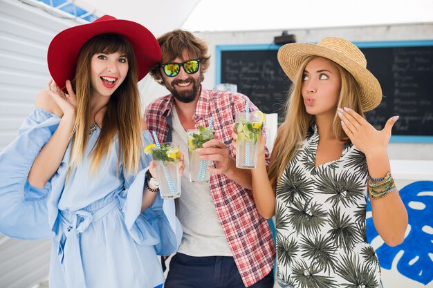 Compañía joven inconformista de amigos de vacaciones en el café de verano, bebiendo cócteles mojito, estilo positivo feliz, sonriendo feliz, dos mujeres y un hombre divirtiéndose juntos, hablando, coqueteando, romance, tres