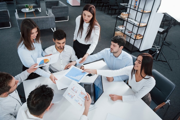 Compartir documentos con gráficos. Vista superior de los trabajadores de oficina en ropa clásica sentados cerca de la mesa usando una computadora portátil y documentos